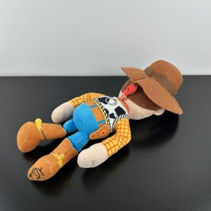 May include: A soft toy of the character Woody from the film Toy Story. Woody is wearing a brown cowboy hat, a yellow plaid shirt, blue trousers, and brown boots. The toy is lying on its side.
