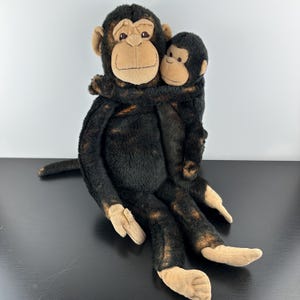 Puede incluir: Un juguete de peluche de mono con un mono más pequeño abrazándolo. Los monos son principalmente negros con caras, manos y pies de color canela. El mono más grande tiene una cola larga y está sentado.