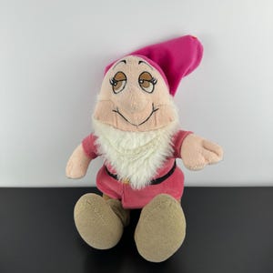 May include: A plush doll of a cartoon character, with a pink hat and outfit, a white beard, and tan skin. The doll has a smiling expression and is sitting with its legs extended. The doll is approximately 25 cm tall.