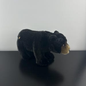 May include: A black plush bear toy with a light brown snout and a small gold detail on its back. The bear is placed on a black surface against a white backdrop. The toy is made of soft, textured material.