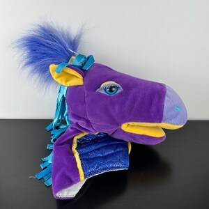 May include: A plush horse head puppet in shades of purple, blue, and yellow. The puppet has a blue mane, yellow ears, and a blue and yellow bridle. The horse's eye is blue with a white outline.