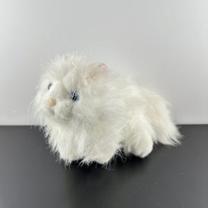 Peut inclure: Chat en peluche blanc avec des yeux bleus. Le chat est assis sur une surface noire.