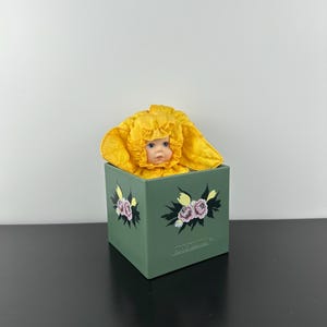 May include: A vintage doll in a yellow flower costume, peeking out of a green box. The box is adorned with floral patterns in pink, yellow, and black. The doll has blue eyes and a calm expression.
