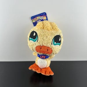 Puede incluir: Un pato de peluche amarillo con patas naranjas y un collar azul con el texto "LPS" en rosa. El pato tiene ojos grandes azules con pupilas negras y un pequeño pico naranja.