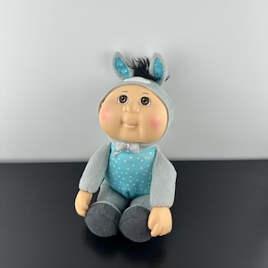 Op de afbeelding: Een grijze pluche Cabbage Patch Kid pop die een blauw en wit gestippeld pak draagt met een grijs ezelkostuum. De pop heeft bruine ogen, roze wangen en een zwarte haardos.