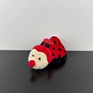 May include: A plush ladybug toy with a red body featuring black spots, a cream-colored face, and a round red nose. The toy has two small black antennae topped with red heart-shaped accents and small black feet visible at the base of the body.