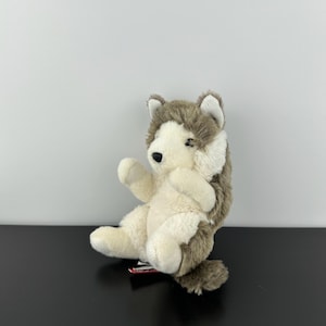 Peut inclure: Une peluche en forme de chien husky assis. Le jouet a un visage et une poitrine blancs, avec de la fourrure grise et brune sur le dos et la tête. Il a un nez noir et est posé sur une surface noire.