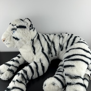 May include: A soft toy white tiger with black stripes. The stuffed animal has blue eyes, a pink nose, and black markings around the eyes and ears. The tiger is lying down, with its legs extended.