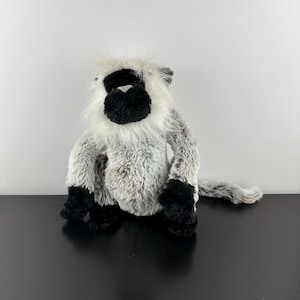 May include: A soft toy monkey with a black face, hands, and feet, and a fluffy white and grey body. The plush animal is sitting on a black surface against a white background.