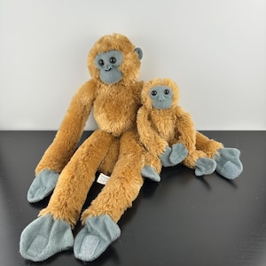 Può includere: Due scimmie di peluche. La scimmia più grande ha braccia e gambe lunghe, con un corpo marrone e piedi e mani grigi. La scimmia più piccola è seduta accanto a quella più grande. Entrambe hanno facce e orecchie grigie.