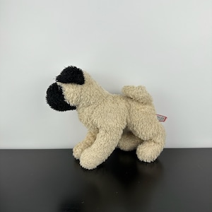 Könnte beinhalten: Ein Stofftier in Form einer Mops-Hunderasse mit beigem Körper und schwarzem Gesicht und Ohren. Das Plüschtier sitzt auf seinen Pfoten, mit einem Etikett an der Seite. Hergestellt aus weichem, flauschigem Material.