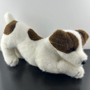 May include: A soft toy dog, white with brown patches and ears, in a playful pose. The dog has a black nose and a fluffy texture. The toy is designed to resemble a real dog, with a friendly appearance.