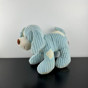 May include: A light blue corduroy dog soft toy with a brown nose and cream-coloured accents. The plush toy has a patchwork design on its body and ears. The dog is standing on a black surface.