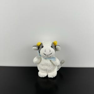May include: A small plush cow toy with white fur, grey patches around the eyes, and yellow horns. The cow features a stitched black mouth and nose, and a light blue ribbon tied around its neck. A white fabric tag is visible on the right side.