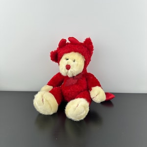 Pode incluir: Um urso de pelúcia vestindo uma fantasia de diabo vermelha com capuz e chifres combinando. O urso tem um rosto, patas e pés cor de creme e um nariz vermelho. O urso está sentado em uma superfície preta.