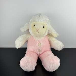 May include: A plush lamb toy with a white head and body, and pink overalls. The lamb has black eyes, a pink nose, and a light yellow face. The overalls are fastened with two beige buttons. The toy is sitting on a black surface.