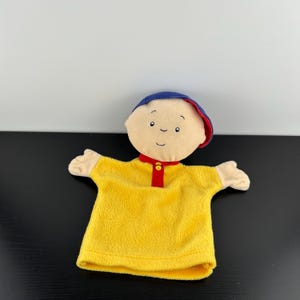 以下が含まれることがあります： 明るいベージュの顔、青い帽子、赤い襟と2つの小さな黄色いボタンが付いた鮮やかな黄色のシャツを着たキャラクターのぬいぐるみハンドパペット。目、眉毛、口などの顔のパーツは刺繍で表現されています。