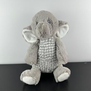 以下が含まれることがあります： 大きな耳と質感のある体を持つ、灰色のぬいぐるみ象。 象の耳の内側と足には白いアクセントがあります。 おもちゃは直立しています。