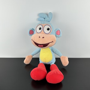 May include: A soft toy monkey with a light blue body, a yellow belly, and red feet. The monkey has large, round eyes, pink ears, and a smiling mouth. The toy is designed to resemble a cartoon character.