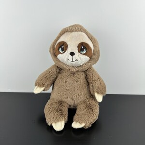 May include: A plush sloth toy with soft brown fur, a lighter beige face, and large, expressive blue eyes. The stuffed animal features a small black embroidered nose and mouth, with cream-coloured claws on its hands and feet, sitting upright.