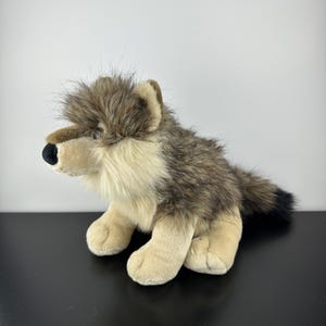 Pode incluir: Um brinquedo de pelúcia de lobo com um design realista. O brinquedo apresenta uma mistura de pele marrom, bege e preta, com um nariz preto e patas bege. O lobo está sentado, com a cabeça virada para a esquerda.