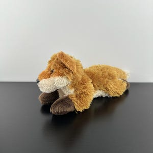 以下が含まれることがあります： ゴールデンブラウンとクリーム色の柔らかい質感の毛並みが特徴のキツネのぬいぐるみ。このぬいぐるみは横たわった姿勢で、ダークブラウンの足、尖った鼻先、小さな立ち耳、黒い鼻を備えており、リアルなデザインで表現されています。