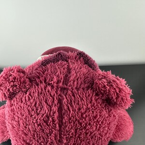 May include: A plush, pink teddy bear with a textured, fuzzy exterior. The bear has a darker pink head and ears, with a lighter pink muzzle. The toy is soft and cuddly, perfect for children or collectors.