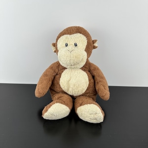 以下が含まれることがあります： 茶色の毛皮とクリーム色の顔、胸、足を持つぬいぐるみのおもちゃの猿。ぬいぐるみには小さな耳と黒いボタンの目があります。猿は暗い表面に直立しています。