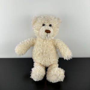 May include: A small, cream-colored teddy bear with a fluffy texture. The bear has a brown nose and small black eyes. Its arms and legs are outstretched, and it is standing upright. The bear is a classic children's toy.