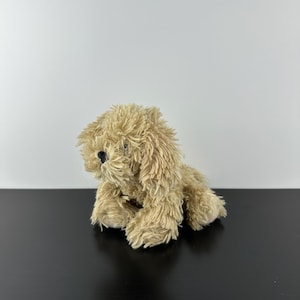 May include: A plush toy dog in a seated position. The dog has soft, shaggy, light brown fur and a black nose. The toy is positioned on a black surface against a white background.