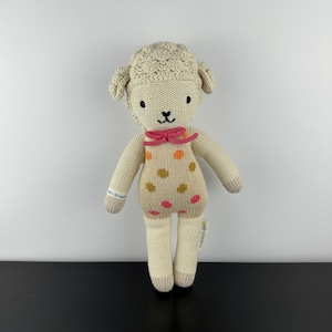 May include: A knitted lamb doll with a cream-colored body, head, and limbs. The lamb has a pink bow tie and a beige romper with orange, pink, and gold polka dots. The doll has black eyes and a black stitched mouth.