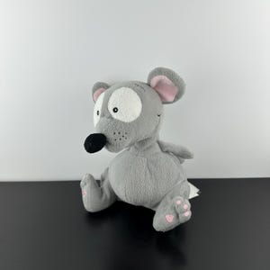 Puede incluir: Un juguete de peluche gris con forma de ratón con grandes ojos blancos, orejas internas rosas y una nariz negra. El ratón tiene almohadillas rosas y está sentado sobre una superficie negra. El juguete es suave y tierno.