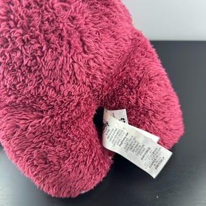 May include: Close-up of a plush toy in a deep pink color. The toy has a soft, fuzzy texture. A white tag with text is attached to the toy. The tag includes the Disney logo and other text.