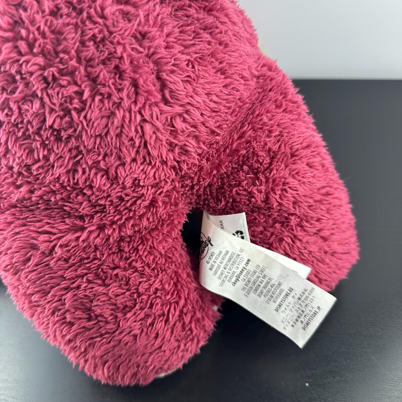 May include: Close-up of a plush toy in a deep pink color. The toy has a soft, fuzzy texture. A white tag with text is attached to the toy. The tag includes the Disney logo and other text.