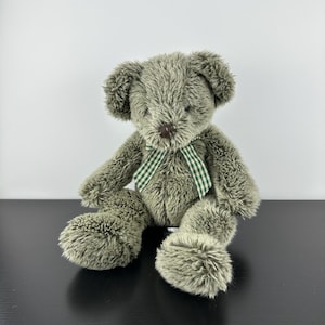 Puede incluir: Un oso de peluche verde oliva con un lazo a cuadros verdes y blancos. El oso tiene una textura suave y peluda y una nariz marrón. Está sentado con los brazos y las piernas extendidos.