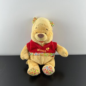 May include: A plush Winnie the Pooh bear wearing a red sweater with a floral pattern and the name "Winnie" embroidered on the front. The bear has a yellow-brown fur and a smiling face.