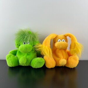 May include: Two plush toys, one green with spiky hair and one yellow with long ears. Both have big eyes and a smiling expression.