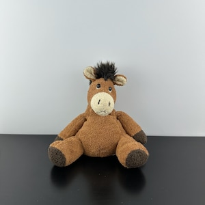 May include: A soft, brown horse toy with a light beige muzzle and ears, dark brown hooves, and a black mane. The plush animal is seated on a black surface against a white backdrop.