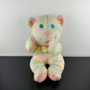 May include: A plush teddy bear toy in pastel colors. The bear has a cream-colored face, pink nose and ears, and blue eyes. Its body is patterned with light green, pink, and yellow plaid. A yellow bow is tied around its neck. The bear is sitting.