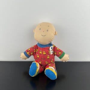以下が含まれることがあります： ハゲ頭のベージュの布製ヘッドとシンプルな刺繍の顔を持つぬいぐるみ。赤い長袖のロンパースを着ており、黄色い鳥の繰り返し模様が特徴で、手首には青い袖口、足には青い布製の靴が縫い付けられています。
