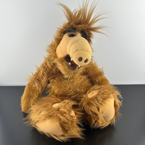 May include: A plush toy character with long, shaggy brown fur, a large tan snout, and dark brown eyebrows. The creature has a tuft of spiky hair on top of its head, a wide open mouth revealing small white teeth, and large tan feet pads.