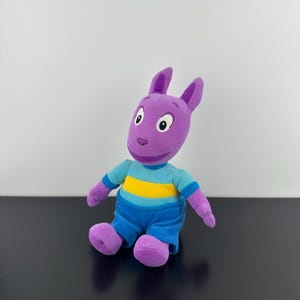 May include: A purple plush kangaroo toy with large ears and embroidered facial features. The stuffed animal wears a light blue shirt with a horizontal yellow stripe across the chest and solid blue pants. The limbs and head are made of purple fabric.