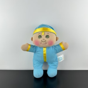 Peut inclure: Une poupée en peluche vêtue d'une combinaison bleue avec des bordures jaunes et un bonnet assorti. La poupée a un visage souriant avec des joues roses et des yeux verts. Une petite étiquette est attachée sur le côté de la poupée.