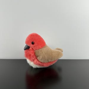 以下が含まれることがあります： 赤い頭と胸、茶色の背中と羽、白いお腹のぬいぐるみ鳥のおもちゃ。鳥は黒い嘴と黒い目を持っています。おもちゃは黒い表面に置かれています。