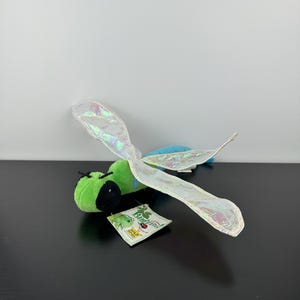 May include: A plush dragonfly toy with green, black, and blue accents. The dragonfly has iridescent wings and a tag that reads "Bugzini". The toy is on a black surface.