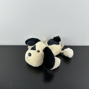 May include: A plush toy dog with cream-colored fur, black ears, and a black spot over one eye. The dog has a small black pom-pom tail and a black pom-pom on its back. The toy is lying on a black surface.