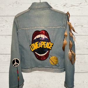 May include: A light blue denim jacket with a red sequined mouth with the words "LOVE & PEACE" in yellow letters. The jacket has a peace sign patch on one sleeve and a brown feather trim on the other sleeve.
