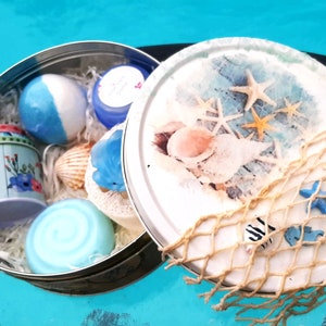 Könnte beinhalten: Eine runde, offene Dose gefüllt mit Badeprodukten und dekorativen Gegenständen. Die Dose enthält eine blau-weiß gestreifte Badekugel, eine kleine Blumentin, Muscheln und eine blaue Seife. Der Deckel zeigt eine Strandszene mit Seesternen und einem dekorativen Netz.