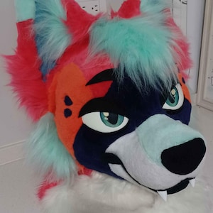May include: A multi-colored faux fur animal head mask featuring bright pink, orange, dark blue, and mint green patches. The mask has large, expressive eyes with mint green irises, a black nose, two white fangs, and a thick white fur neck ruff.