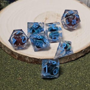 May include: A collection of seven polyhedral dice, each with a clear resin body and blue numbering. The dice feature internal inclusions of dark red and teal elements. The dice are arranged on a wooden slice and green surface.